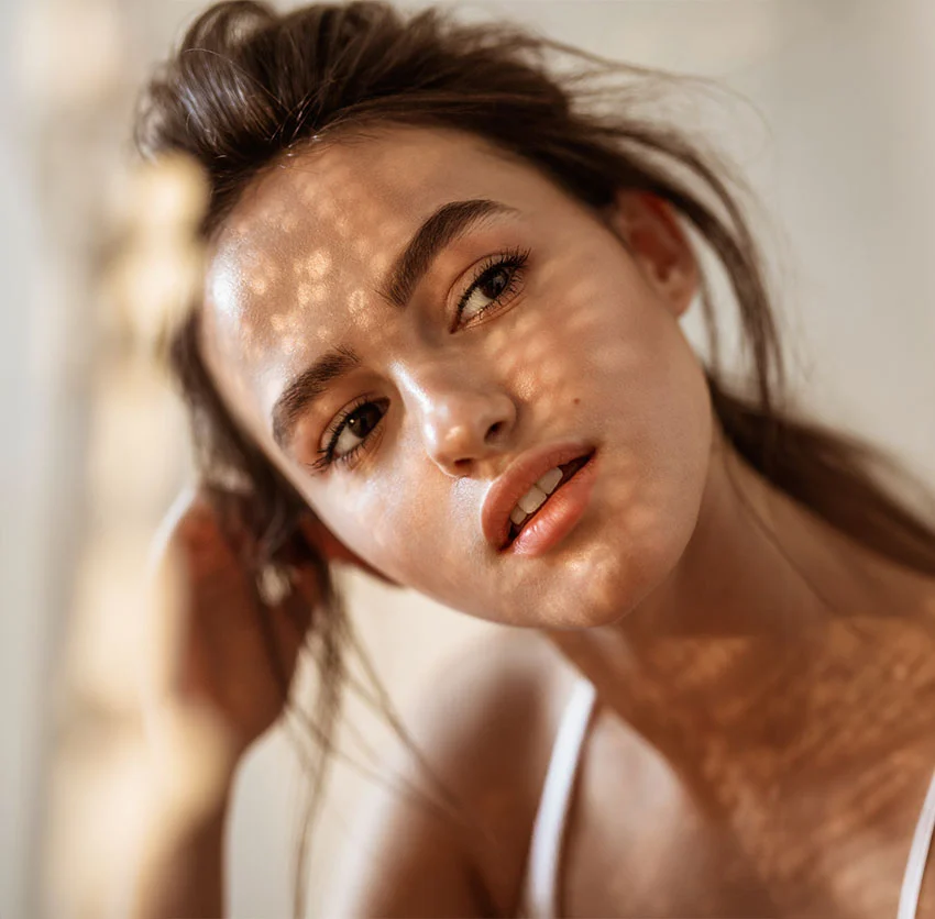 A young woman with her hair loosely tied back, gazes thoughtfully to the side as sunlight creates a dappled pattern on her face - Acne Treatment in Chino, CA