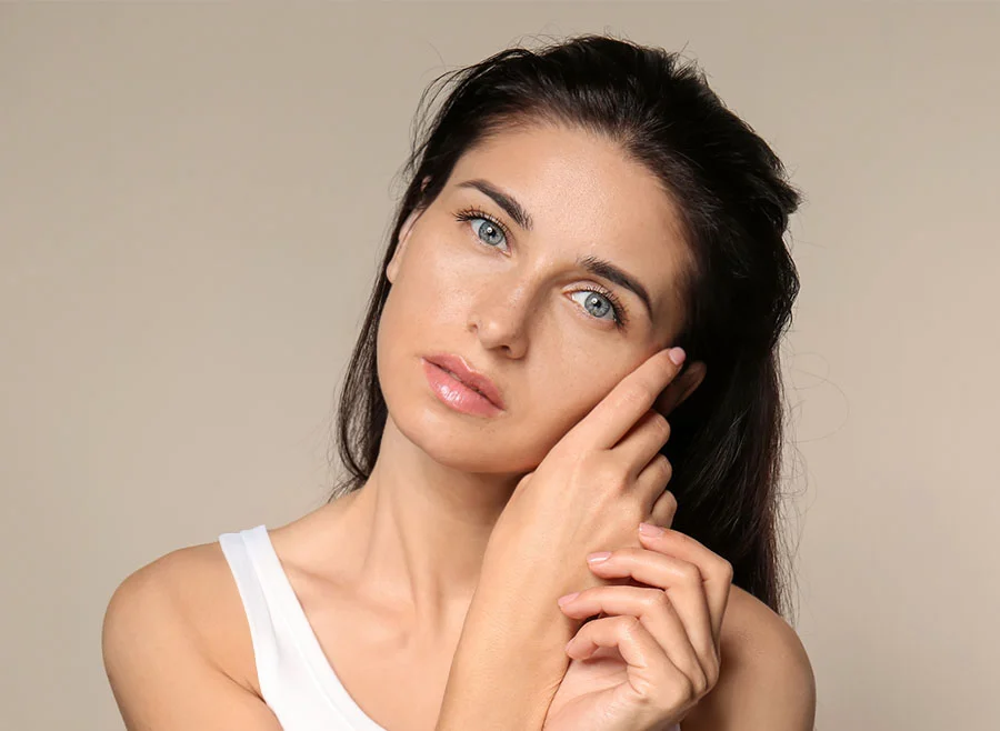 A woman with long dark hair and piercing blue eyes gently touches her face with both hands, wearing a white tank top against a neutral background - Acne Treatment in Chino, CA