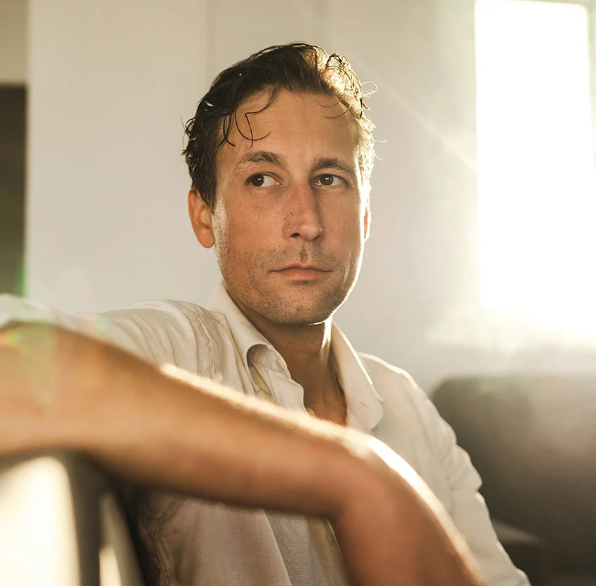 A man with short hair, wearing a white shirt, sits pensively in a sunlit room with light streaming in through a window - Biote Bioidentical Hormone Treatment in Chino, CA
