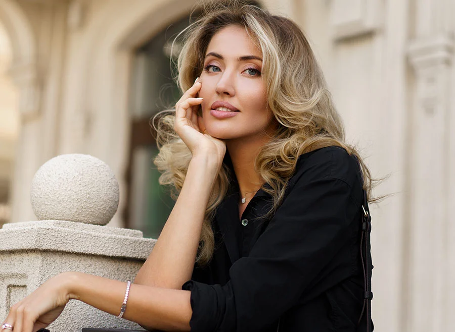 A woman with long blonde hair, wearing a black outfit, leans on an outdoor ledge, smiling gently - Biote Bioidentical Hormone Treatment in Chino, CA