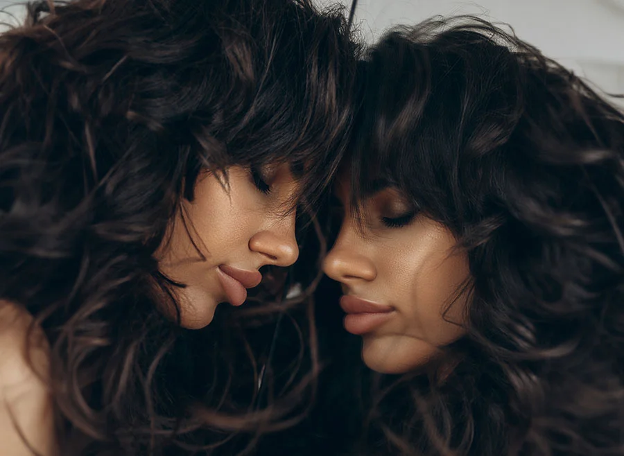 A woman with voluminous dark hair looks down with her eyes closed, her face mirrored closely beside her reflection - Cheek Filler in Chino, CA