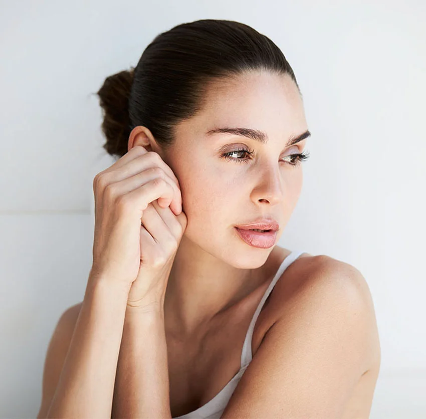 A woman with dark hair tied back, gently touching her ear, looks to the side with a serene expression, wearing a white tank top against a light background - Chemical Peels in Chino, CA