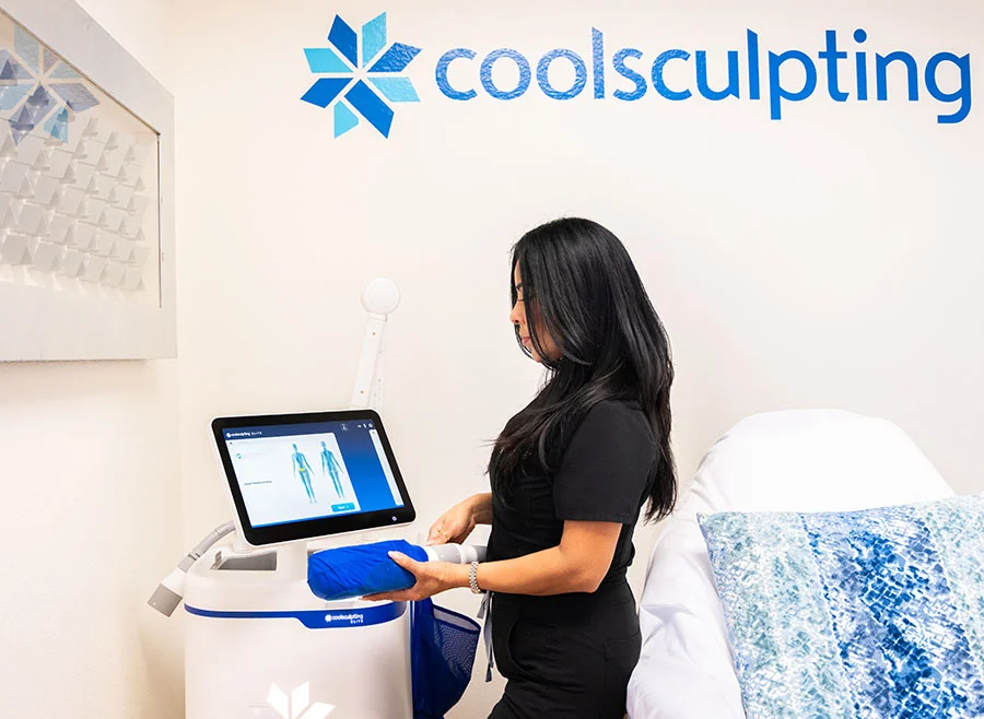 A woman in a black uniform operates a CoolSculpting machine in a clinical setting with the CoolSculpting logo on the wall behind her - Coolsculpting in Chino, CA
