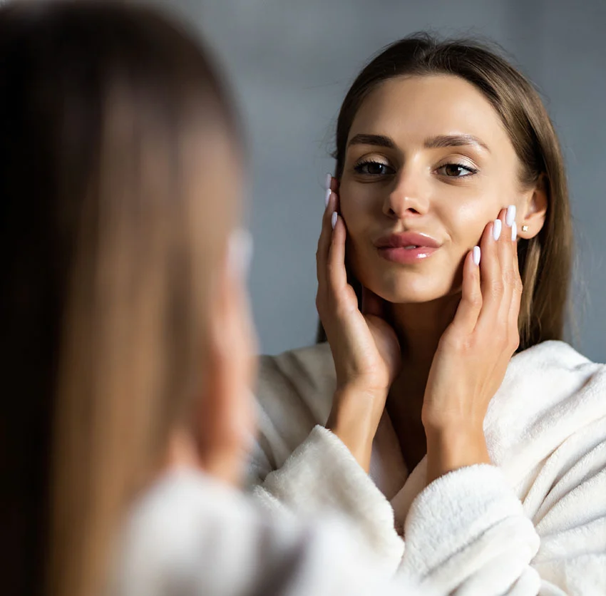 A woman in a white robe gently touches her face while looking at herself in the mirror, smiling softly - Dermal Fillers in Chino, CA