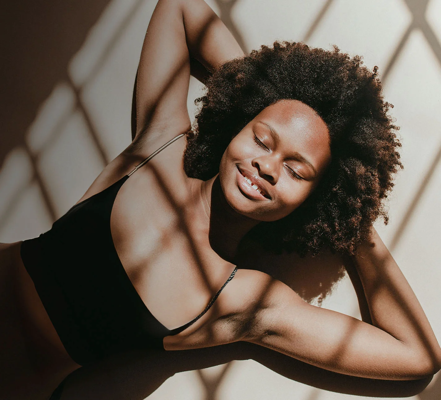 A woman with dark curly hair, wearing a black tank top, lying in a dim room with shadows falling over her - Health and Wellness in Chino, CA