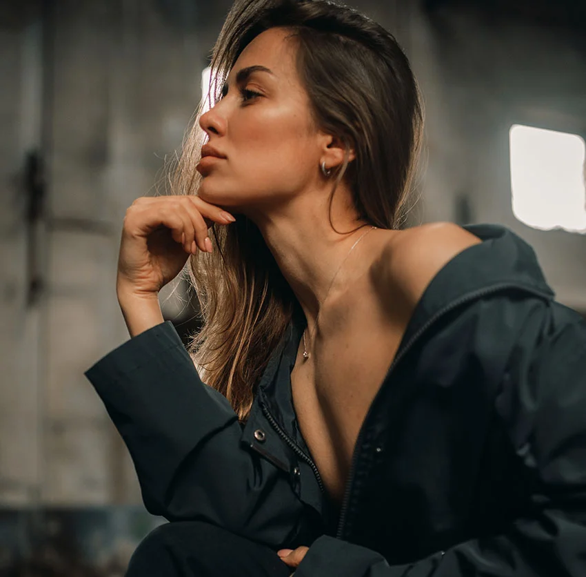 A woman with long hair sits thoughtfully, resting her chin on her hand, wearing a dark jacket, in an industrial setting - Kybella in Chino, CA