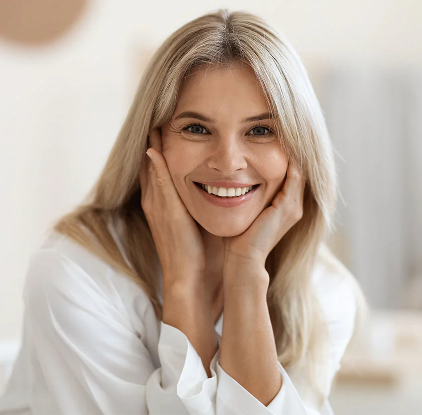 A woman with blonde hair and a white robe smiles warmly, with hands touching her face, in a bright room - Liquid Facelift in Chino, CA