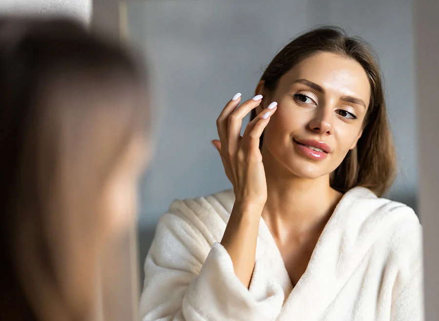A woman with a white robe applies skincare product to her face while looking in a mirror - Liquid Facelift in Chino, CA