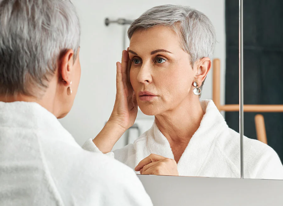 A woman with short gray hair examines her face in a bathroom mirror, looking thoughtful - Mint Thread Facelift in Chino, CA